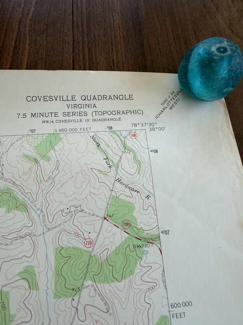 May include: A topographic map titled COVESVILLE QUADRANGLE VIRGINIA 7.5 MINUTE SERIES with contour lines, green forested areas, and road markings, accompanied by a smooth, rounded, translucent teal glass object resting on the upper right corner.