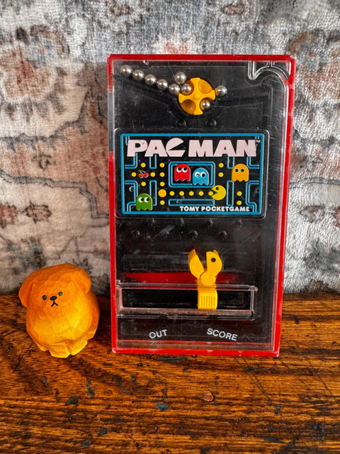 May include: A vintage Tomy Pocketgame Pac-Man handheld game featuring a black and red plastic casing with a maze graphic, metal ball bearings, and a yellow character controller, alongside a small, round, orange carved wooden dog figurine.
