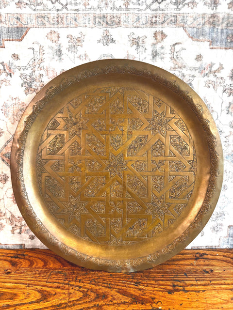 May include: A round brass tray featuring an intricate engraved geometric star pattern in the center and a decorative border along the rim. The metal surface has a warm, aged patina with detailed line work throughout the central design.