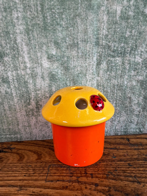 May include: A ceramic mushroom-shaped flower frog with a bright orange cylindrical base and a glossy yellow cap. The cap features several circular holes for flower stems and a small, three-dimensional red ladybug with black spots attached to the top.