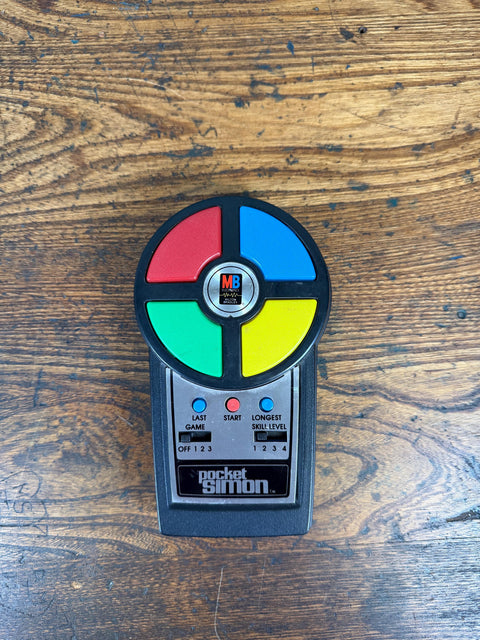 May include: A black handheld electronic game featuring a circular top with four large red, blue, green, and yellow buttons. The center displays an MB Milton Bradley logo. Below are three small buttons labeled Last Game, Start, and Longest Skill Level, with two sliding switches and the text pocket simon.
