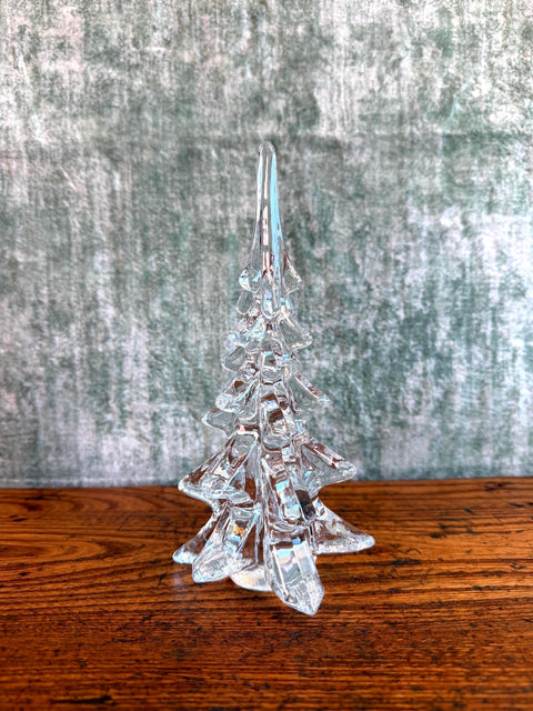 May include: A clear glass figurine shaped like a pine tree with a pointed top and tiered, angular branches. The glass is transparent and smooth, reflecting light across its faceted surface to create a crystalline appearance.