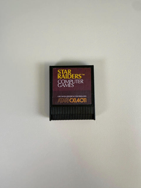 May include: A black Atari Star Raiders computer game cartridge featuring a dark purple label with yellow text that reads Star Raiders, Computer Games, Use With Joystick Controllers, and Atari CXL4011.