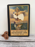 May include: An antique copy of "The Adventures of Huckleberry Finn" by Mark Twain, featuring a boy on a log illustration. A small brown dog figurine rests in front of the book on a white surface.