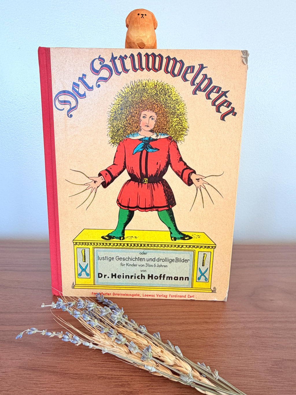 May include: A vintage copy of "Der Struwwelpeter" with a wooden dog figurine on top. The book cover features a colorful illustration of a figure in a red coat and green tights. The title is in large, ornate letters.
