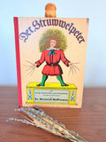 May include: A vintage copy of "Der Struwwelpeter" with a wooden dog figurine on top. The book cover features a colorful illustration of a figure in a red coat and green tights. The title is in large, ornate letters.