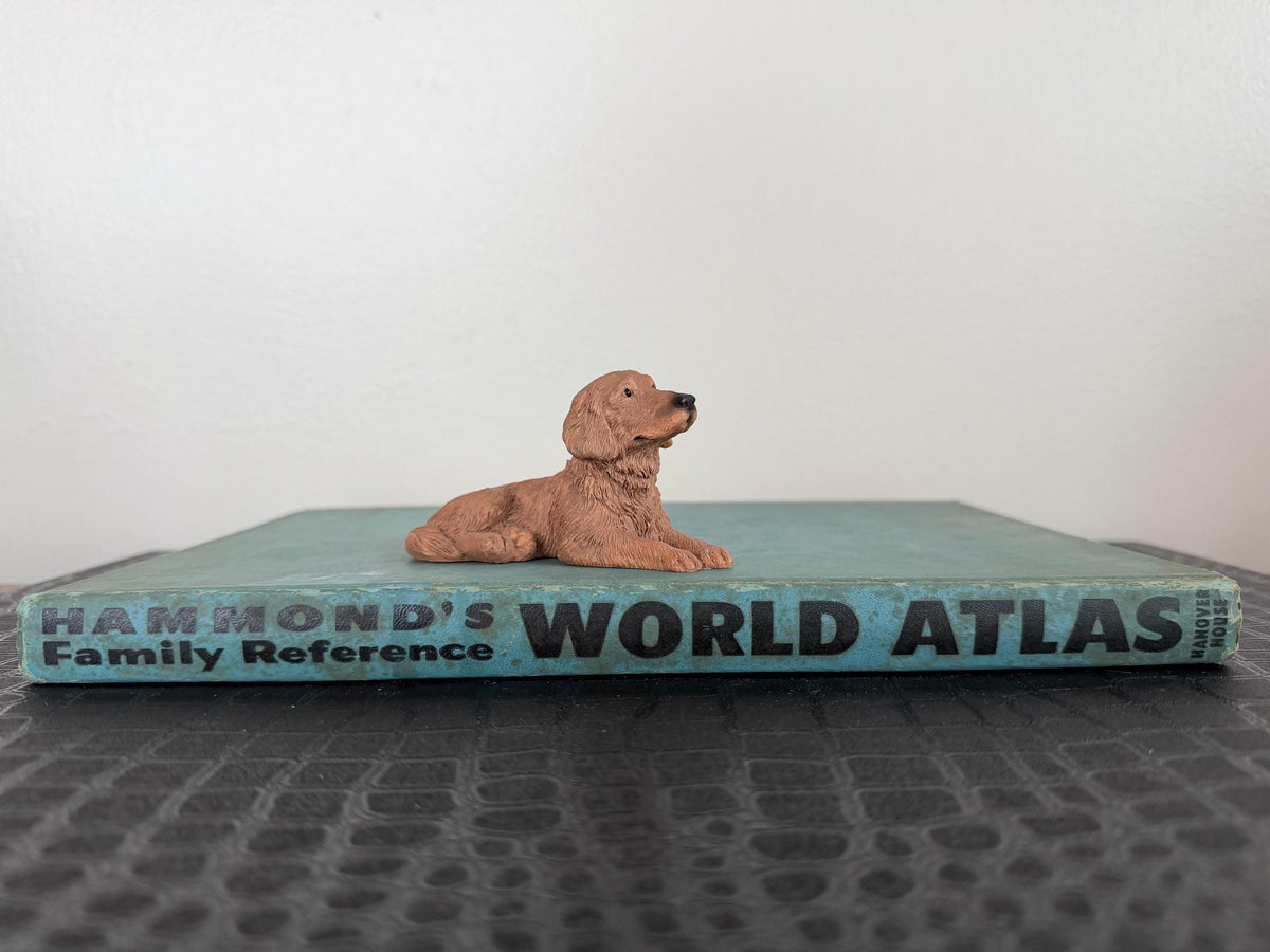 May include: A small, brown dog figurine rests on a vintage blue "WORLD ATLAS" book. The book's cover features the text "HAMMOND'S FAMILY REFERENCE". The dog is lying down, facing right, with its head raised.
