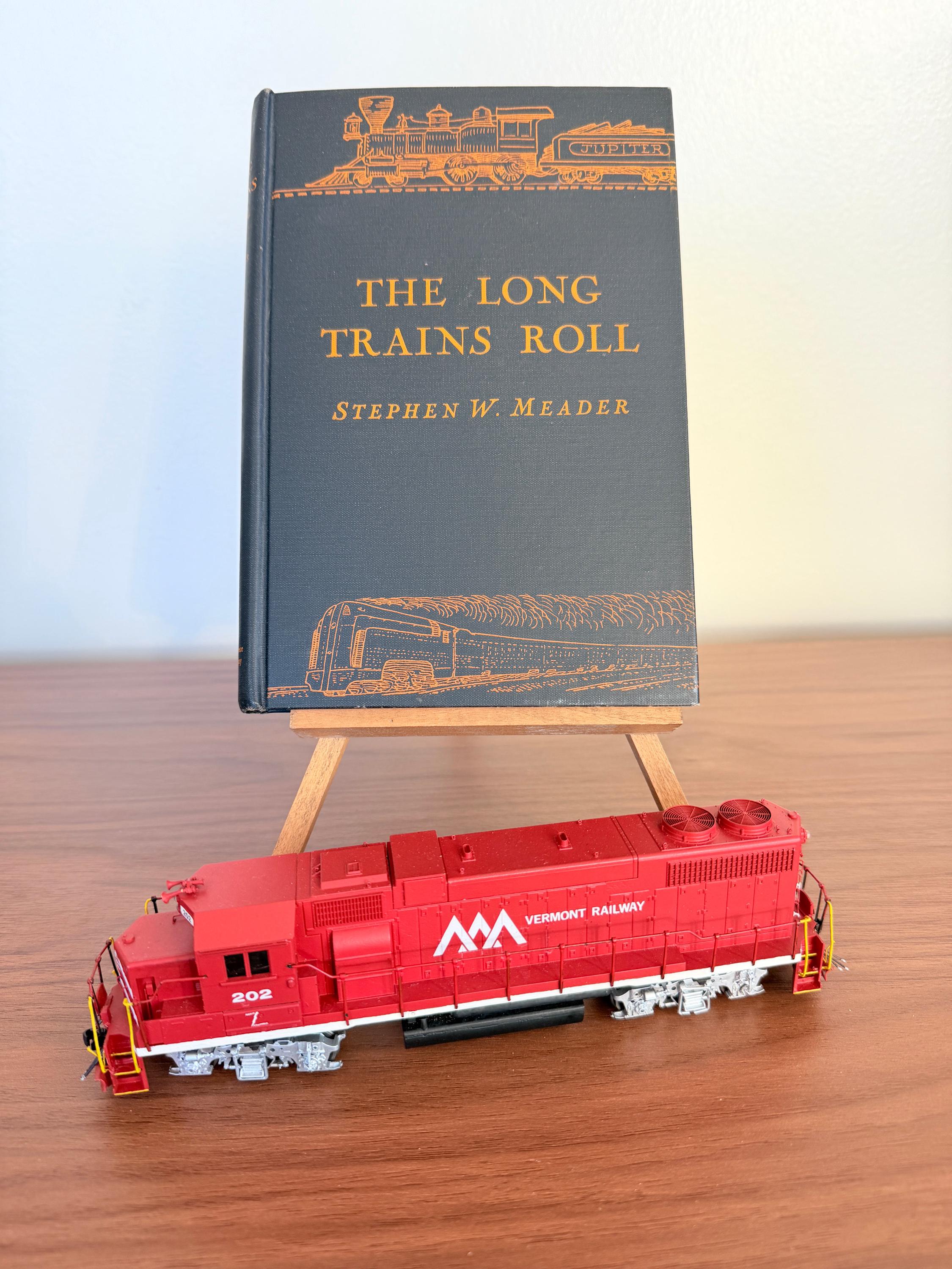 May include: A red Vermont Railway model train sits in front of a book titled "The Long Trains Roll" by Stephen W. Meader. The book is propped up on a small wooden easel.