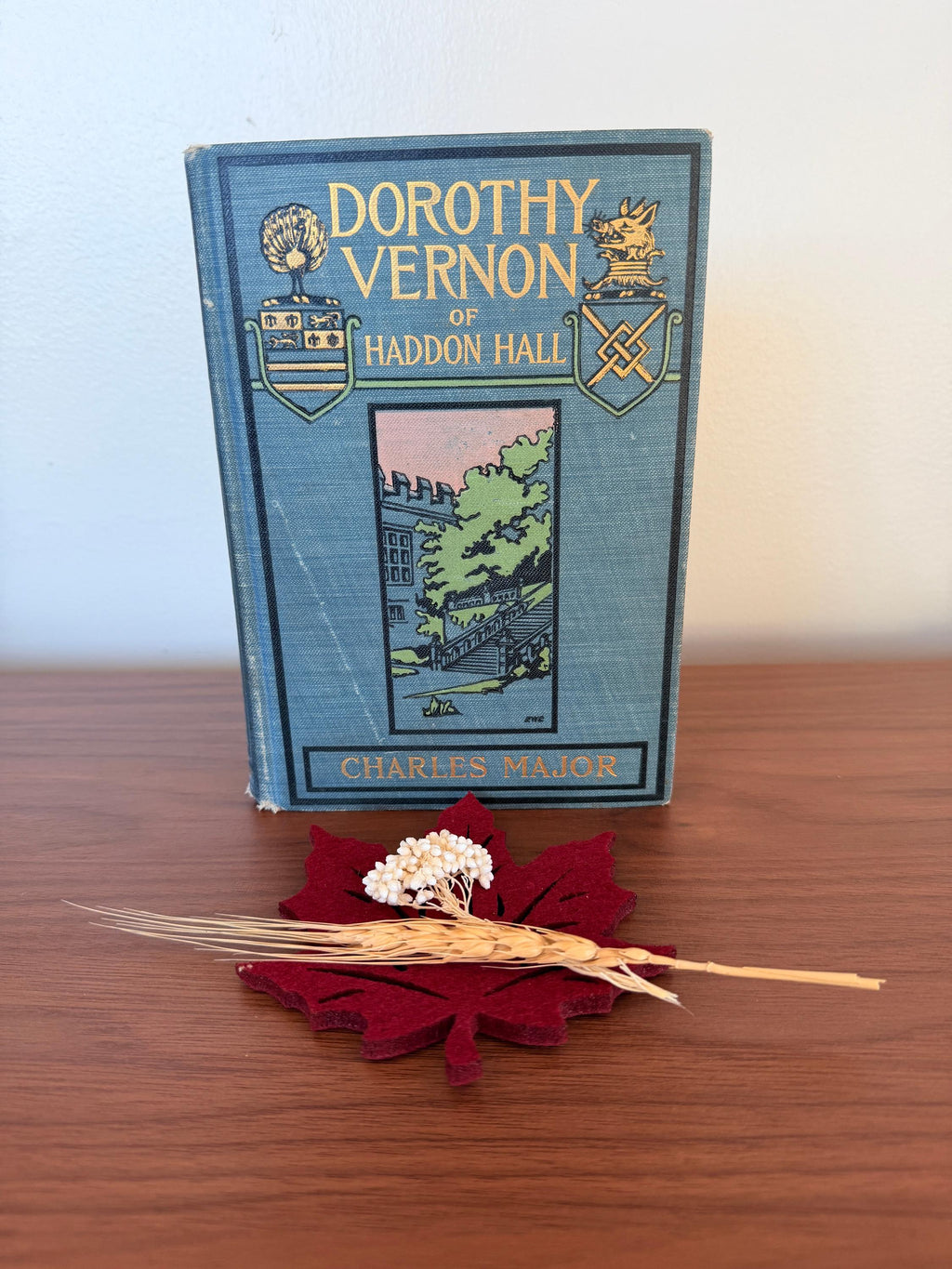 May include: A vintage book titled "Dorothy Vernon of Haddon Hall" with a blue cover and gold lettering. A burgundy leaf-shaped felt coaster holds a sprig of white flowers and a wheat stalk. The book is on a wooden surface.