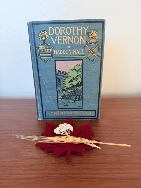 May include: A vintage book titled "Dorothy Vernon of Haddon Hall" with a blue cover and gold lettering. A burgundy leaf-shaped felt coaster holds a sprig of white flowers and a wheat stalk. The book is on a wooden surface.