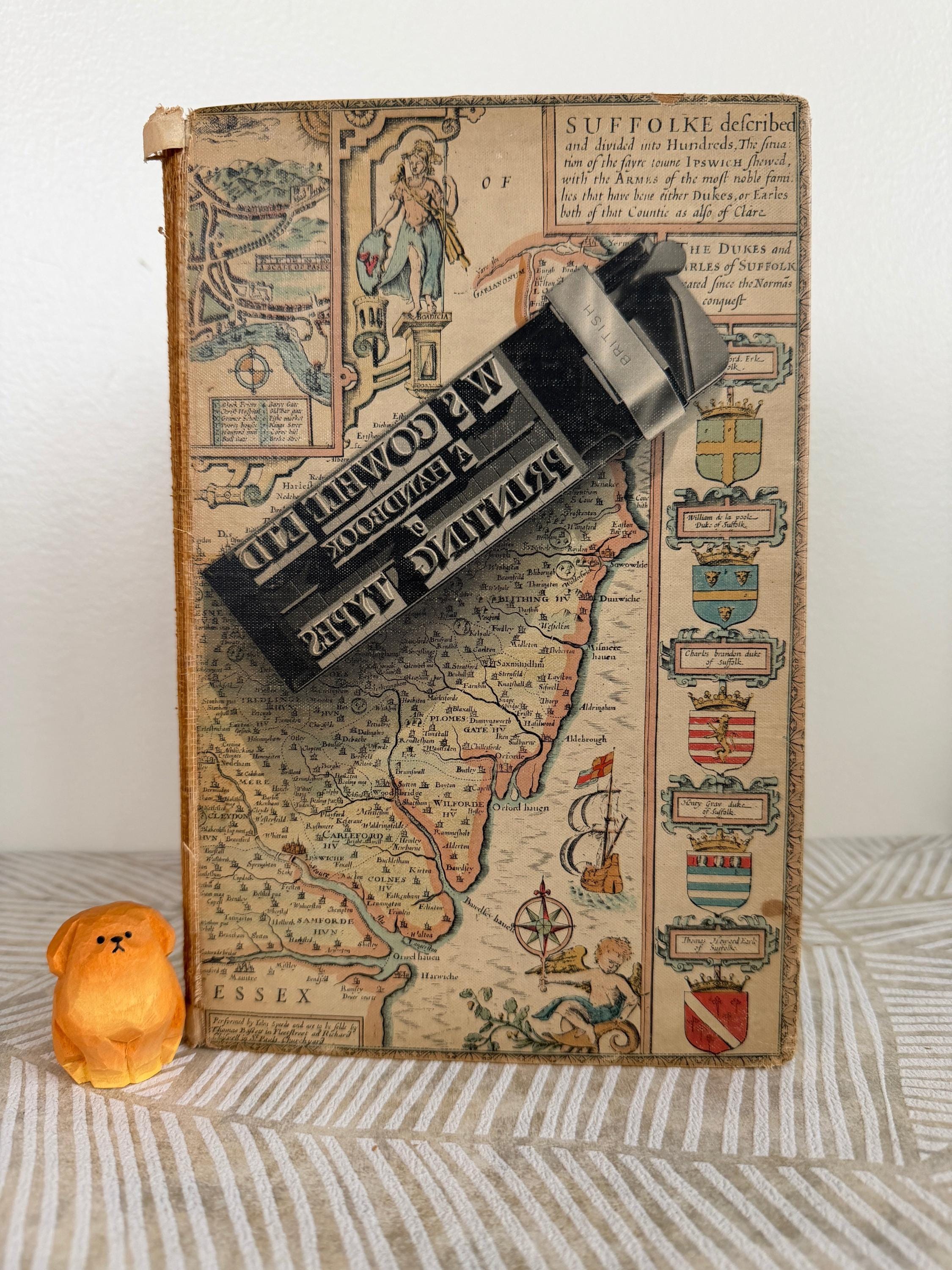 May include: An antique book with a detailed map illustration, featuring text and decorative elements. A black printing press tool rests on the book. A small, orange, dog-shaped figurine sits nearby. The book's cover displays historical cartography.