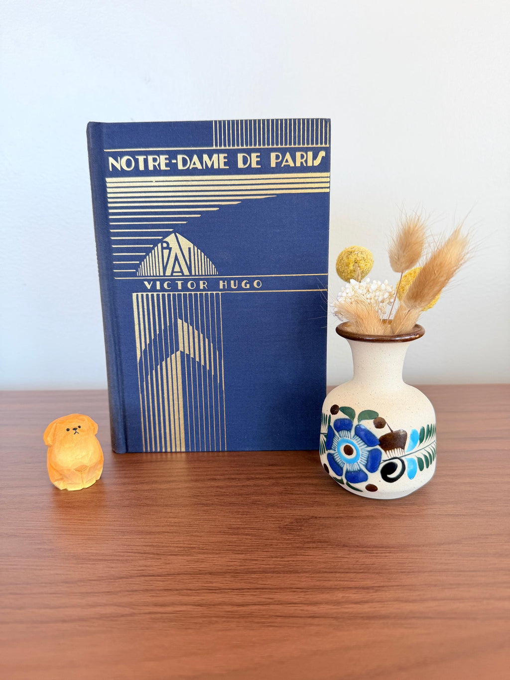 May include: A still life arrangement featuring a vintage book titled "Notre-Dame de Paris" by Victor Hugo, a small ceramic vase with blue floral designs, and a small orange dog figurine. The vase holds dried flowers.