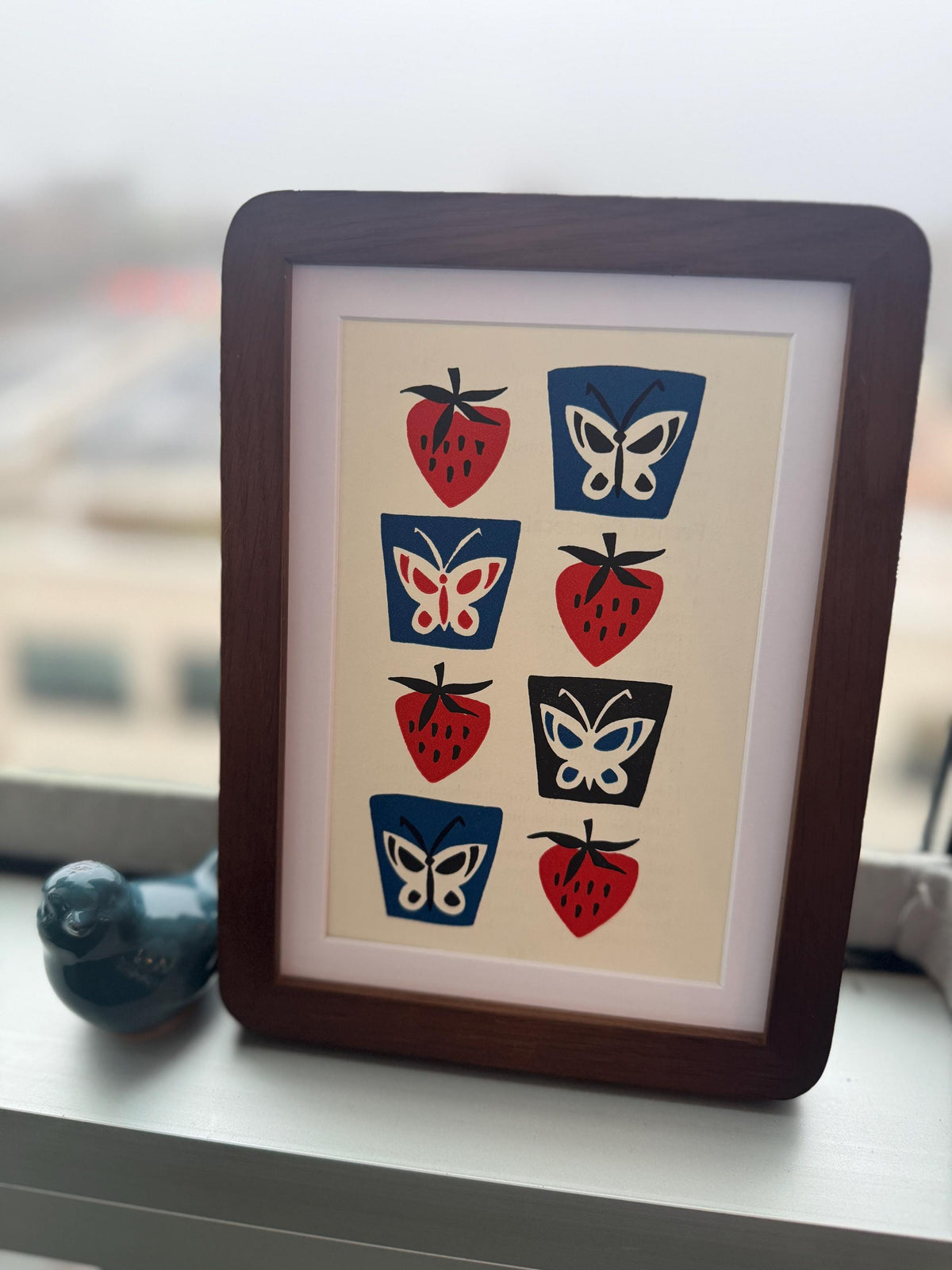 May include: Framed art print featuring a repeating pattern of red strawberries and blue, black, and white butterflies. The artwork is set against a cream background and framed in a dark brown wooden frame. A decorative blue bird figurine sits to the left.