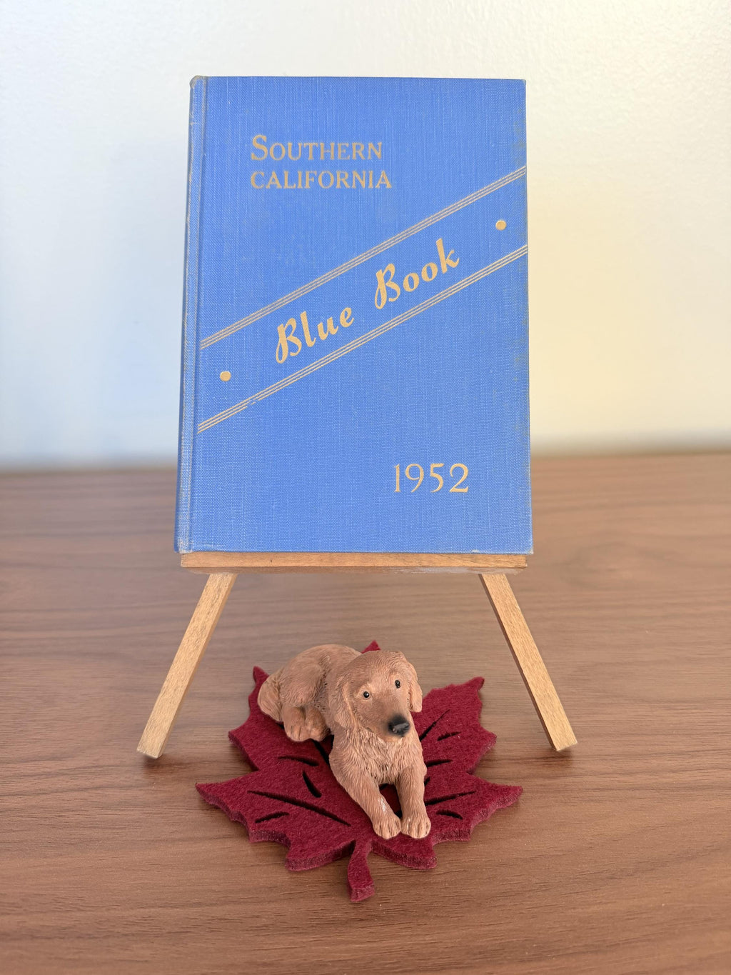 May include: A vintage blue "Southern California Blue Book" from 1952, displayed on a small wooden easel. A brown dog figurine rests on a red felt leaf in front of the book. The book's title is in gold lettering.
