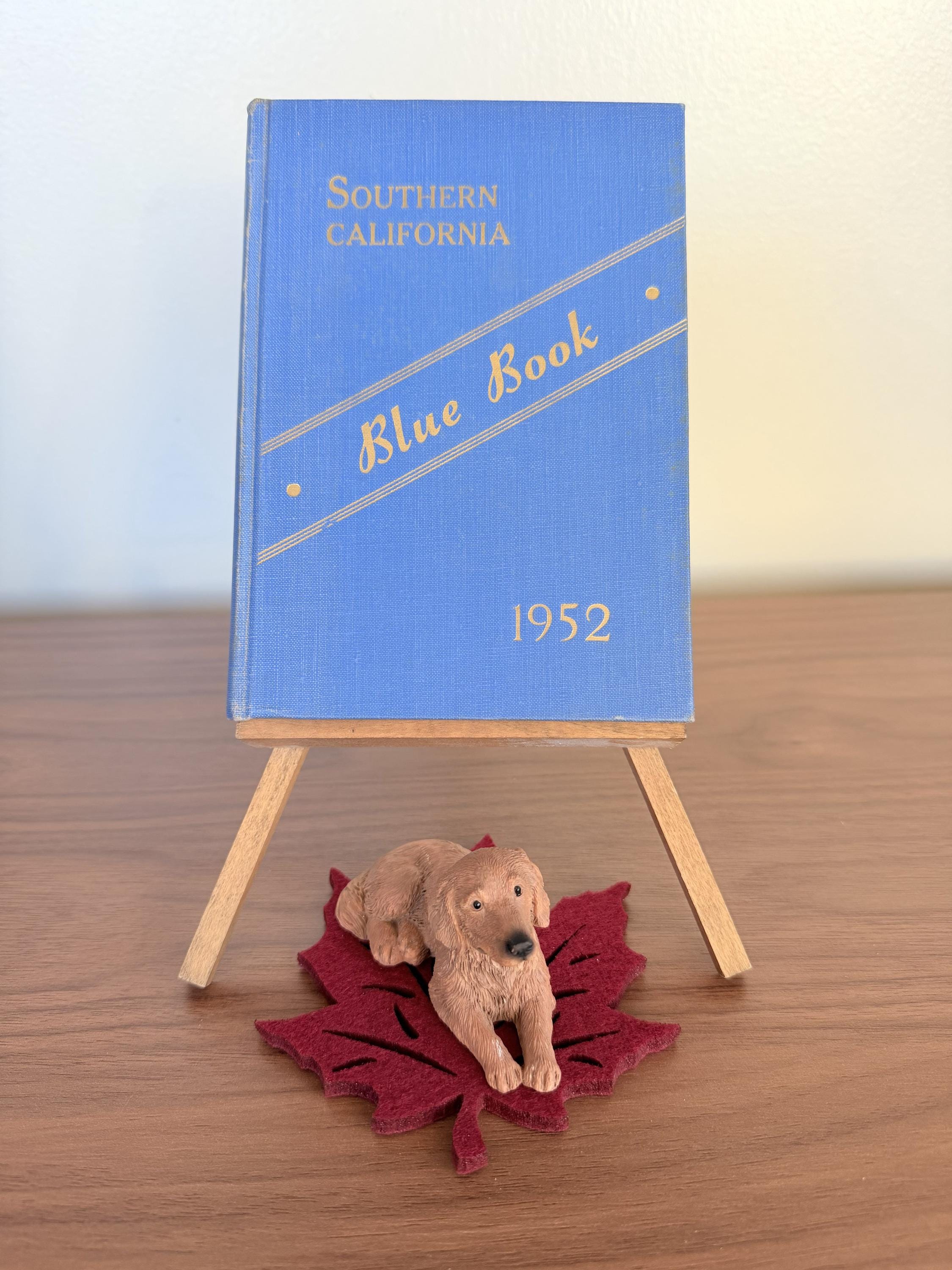 May include: A vintage blue "Southern California Blue Book" from 1952, displayed on a small wooden easel. A brown dog figurine rests on a red felt leaf in front of the book. The book's title is in gold lettering.