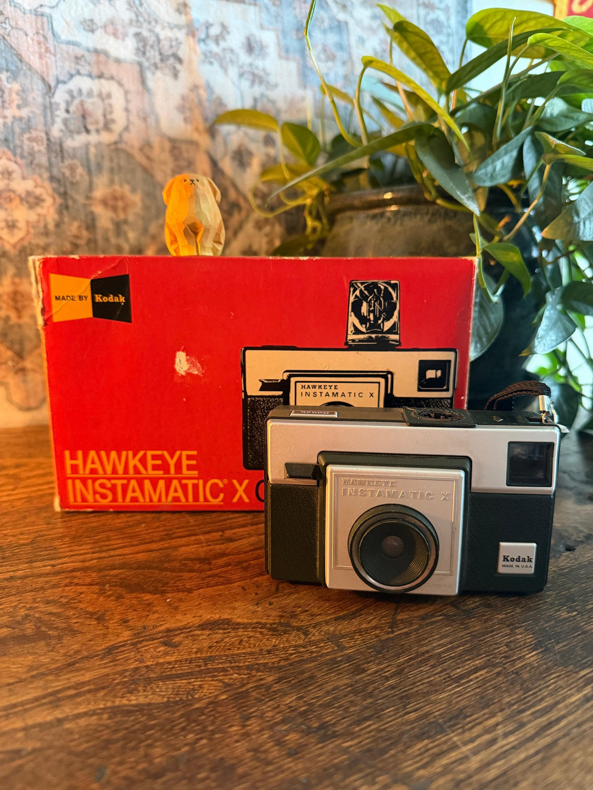 May include: A vintage Kodak Hawkeye Instamatic X camera with its original red box. The camera is silver and black, with the Kodak logo visible. The box has the text "Hawkeye Instamatic X" and "Made by Kodak". A small wooden dog figurine sits on top of the box.