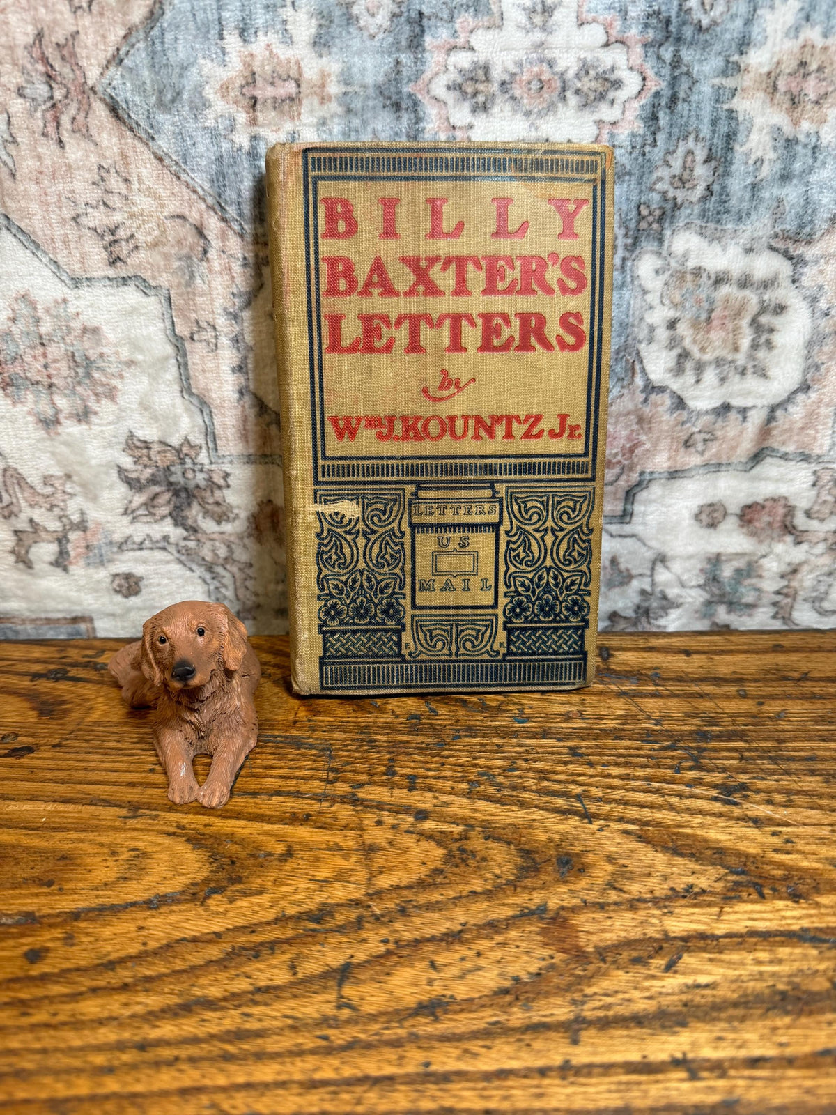 May include: A vintage book titled "Billy Baxter's Letters" with red and black text on a tan cover. A small brown dog figurine rests beside the book on a wooden surface. The book's cover features an illustration of a mailbox.