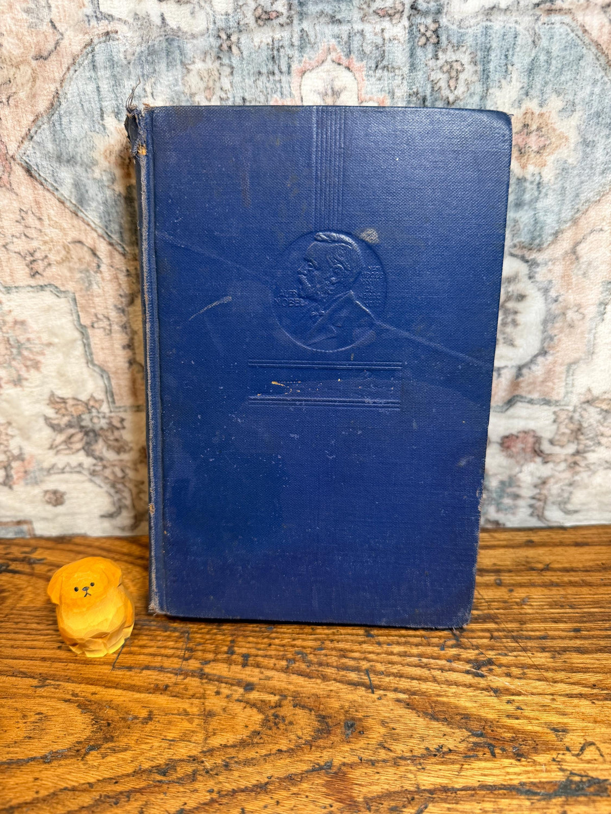 May include: A vintage, navy blue book with a portrait on the cover and a small, carved wooden dog figurine. The book's cover shows signs of wear. The figurine is a light yellow color and is sitting on a wooden surface.