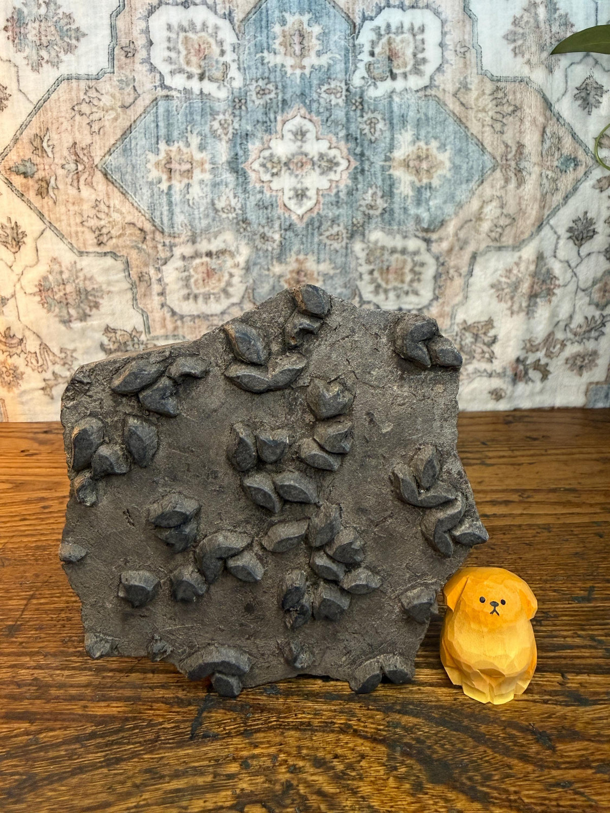 May include: A dark gray, rectangular stone block with carved leaf-like shapes, next to a small, carved wooden dog figurine. The dog is light orange and has a simple, stylized design. The background features a patterned rug.