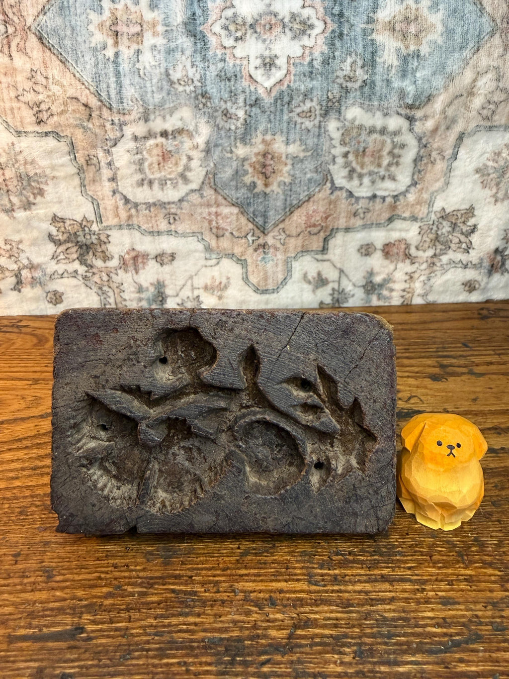 May include: A dark brown, rectangular wooden mold with carved floral designs and a small, yellow wooden dog figurine. The mold has a rough, textured surface, and the dog is intricately carved.