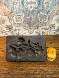 May include: A dark brown, rectangular wooden mold with carved floral designs and a small, yellow wooden dog figurine. The mold has a rough, textured surface, and the dog is intricately carved.