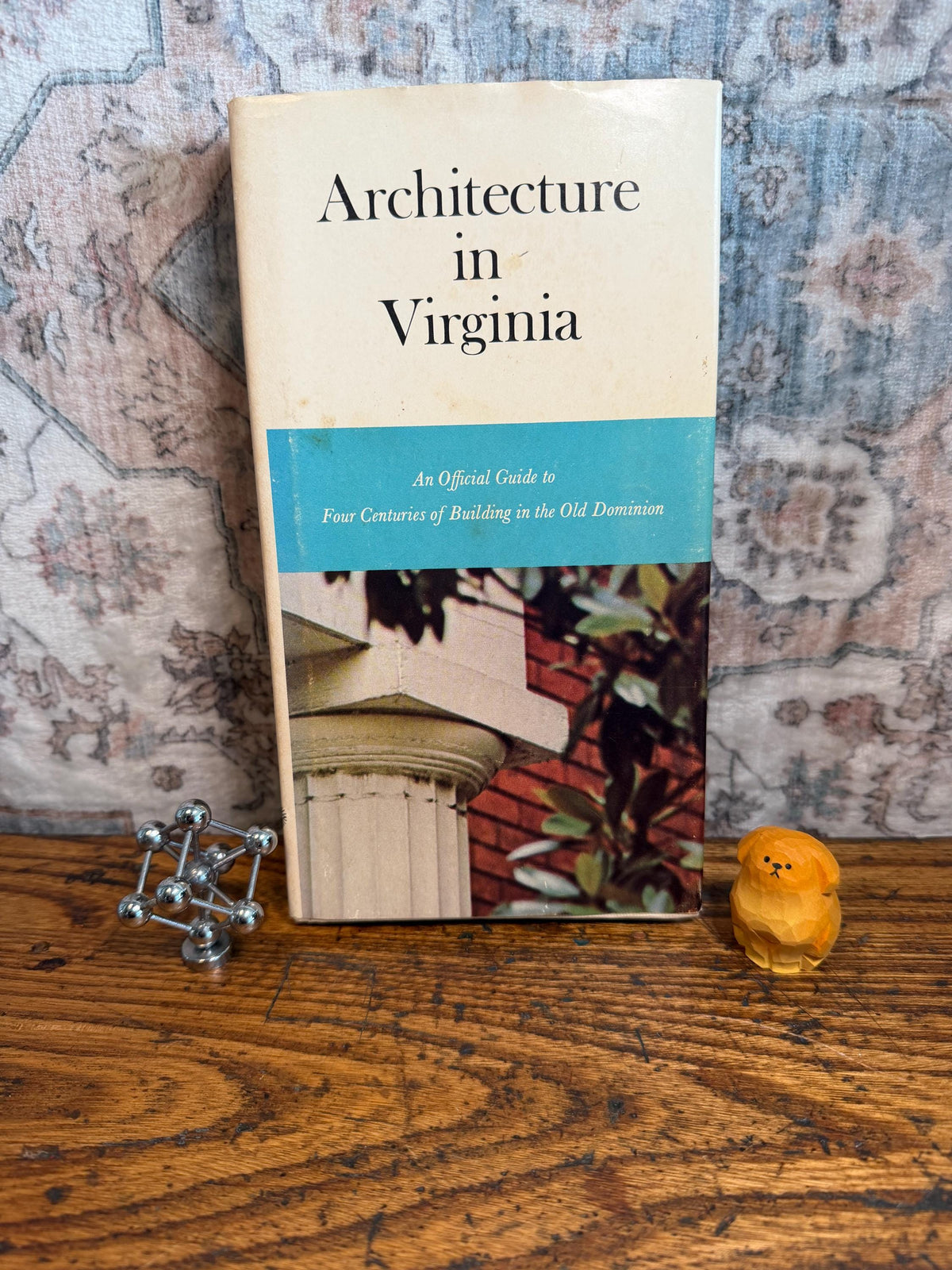 May include: A vintage book titled "Architecture in Virginia" with a blue stripe, accompanied by a silver geometric sculpture and a small wooden dog figurine. The book cover features architectural details.
