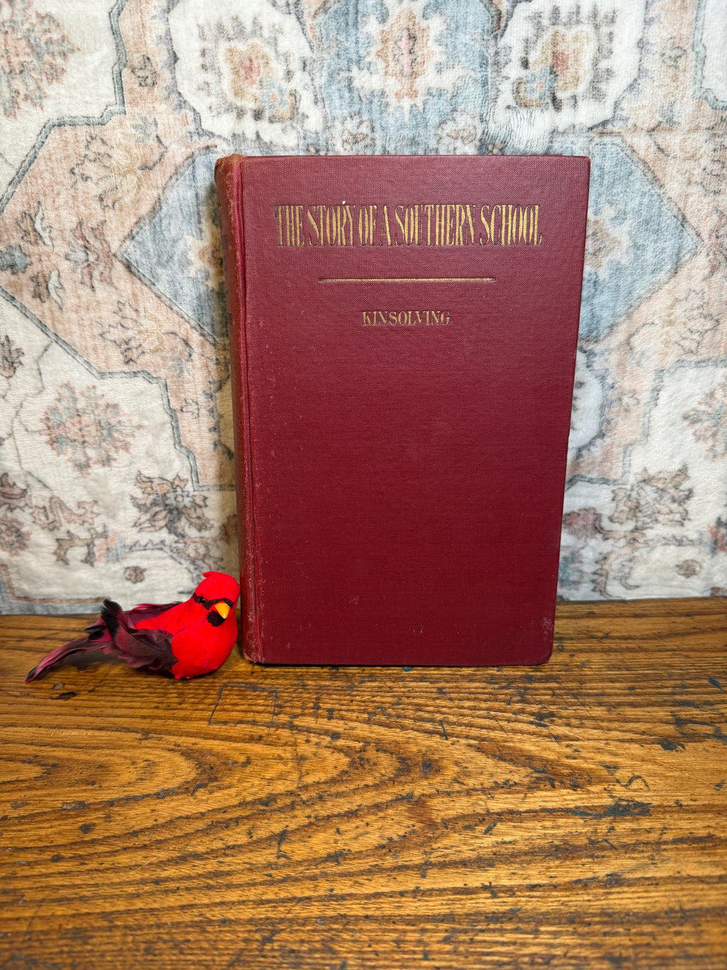 May include: A maroon-colored book titled "THE STORY OF A SOUTHERN SCHOOL" with gold lettering. The author's name, KINSOLVING, is below the title. A red decorative bird sits to the left of the book on a wooden surface.