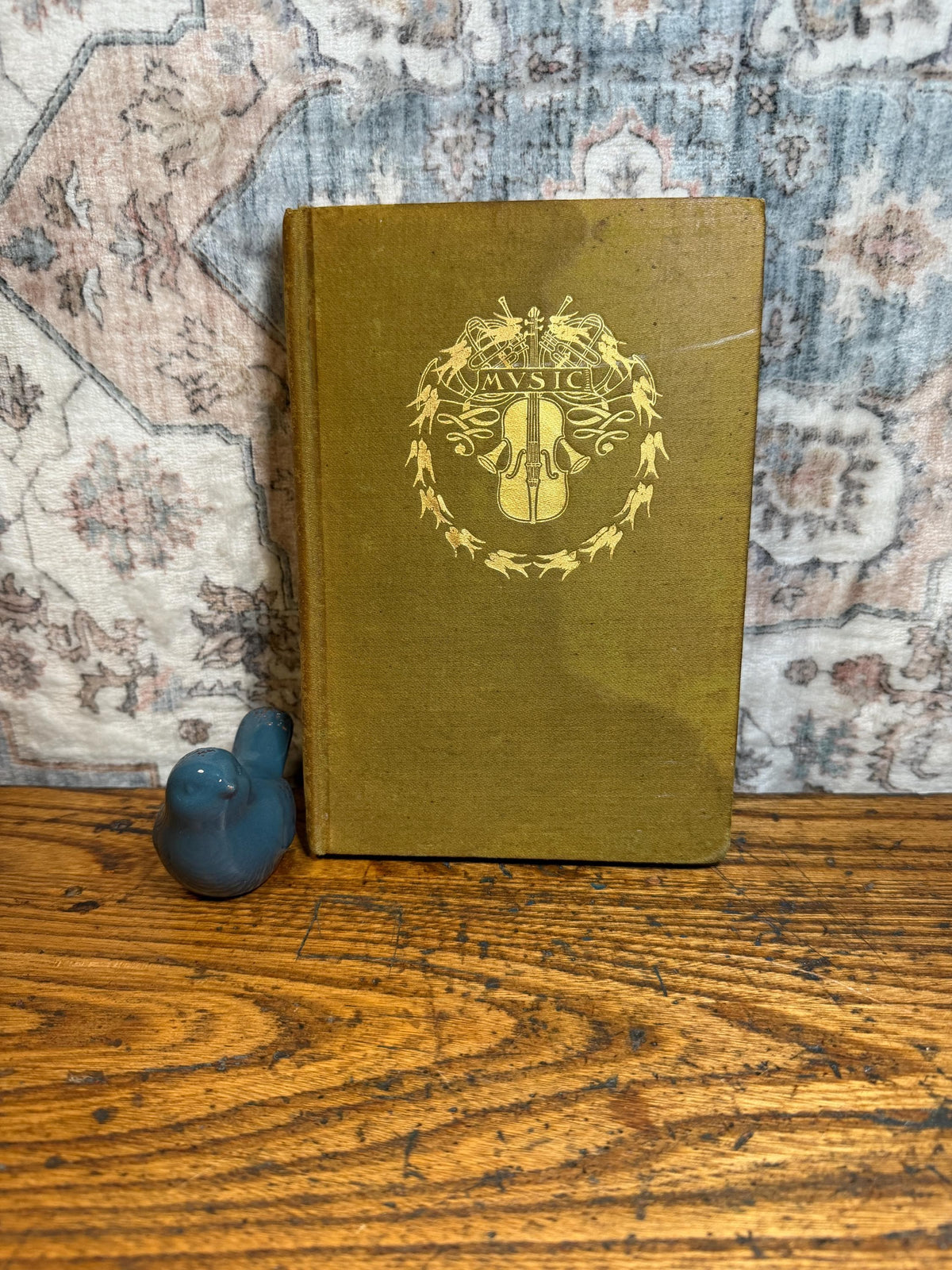 May include: A vintage book with a gold-colored cover featuring a gold emblem of a violin and the word "MUSIC". A small blue bird figurine sits next to the book on a wooden surface.