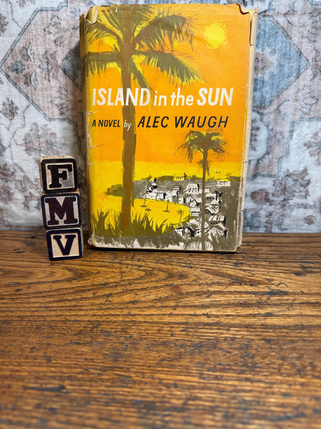 May include: A vintage book titled "ISLAND in the SUN" by Alec Waugh, featuring a yellow cover with a palm tree illustration. The book is set on a wooden surface, with wooden letter blocks spelling "FMV" in the foreground.