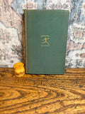 May include: A vintage green book with a gold emblem of a person running is next to a small, carved wooden dog figurine. The book and figurine are on a wooden surface with a patterned rug in the background.