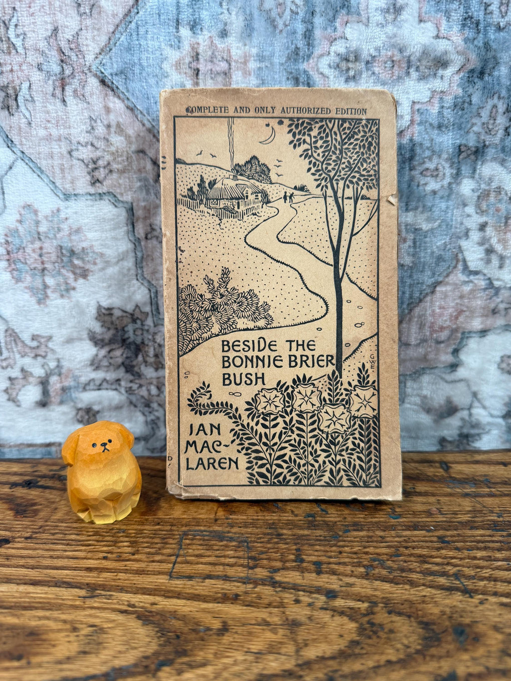 May include: A vintage book titled Beside the Bonnie Brier Bush by Ian Maclaren with a tan cover featuring a black ink illustration of a cottage, a winding path, and trees, accompanied by a small, hand-carved yellow wooden dog figurine.