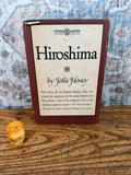May include: A hardcover book titled Hiroshima by John Hersey with a dark red spine and border, featuring a cream-colored cover with black text and a small yellow carved wooden dog figurine positioned to the left of the book.