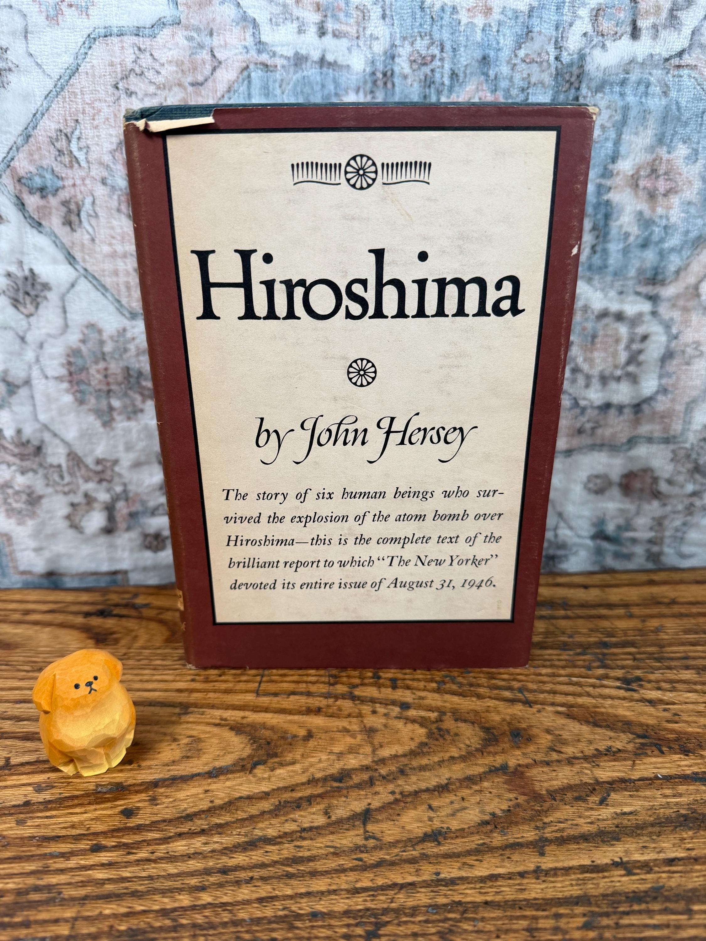 May include: A hardcover book titled Hiroshima by John Hersey with a dark red spine and border, featuring a cream-colored cover with black text and a small yellow carved wooden dog figurine positioned to the left of the book.