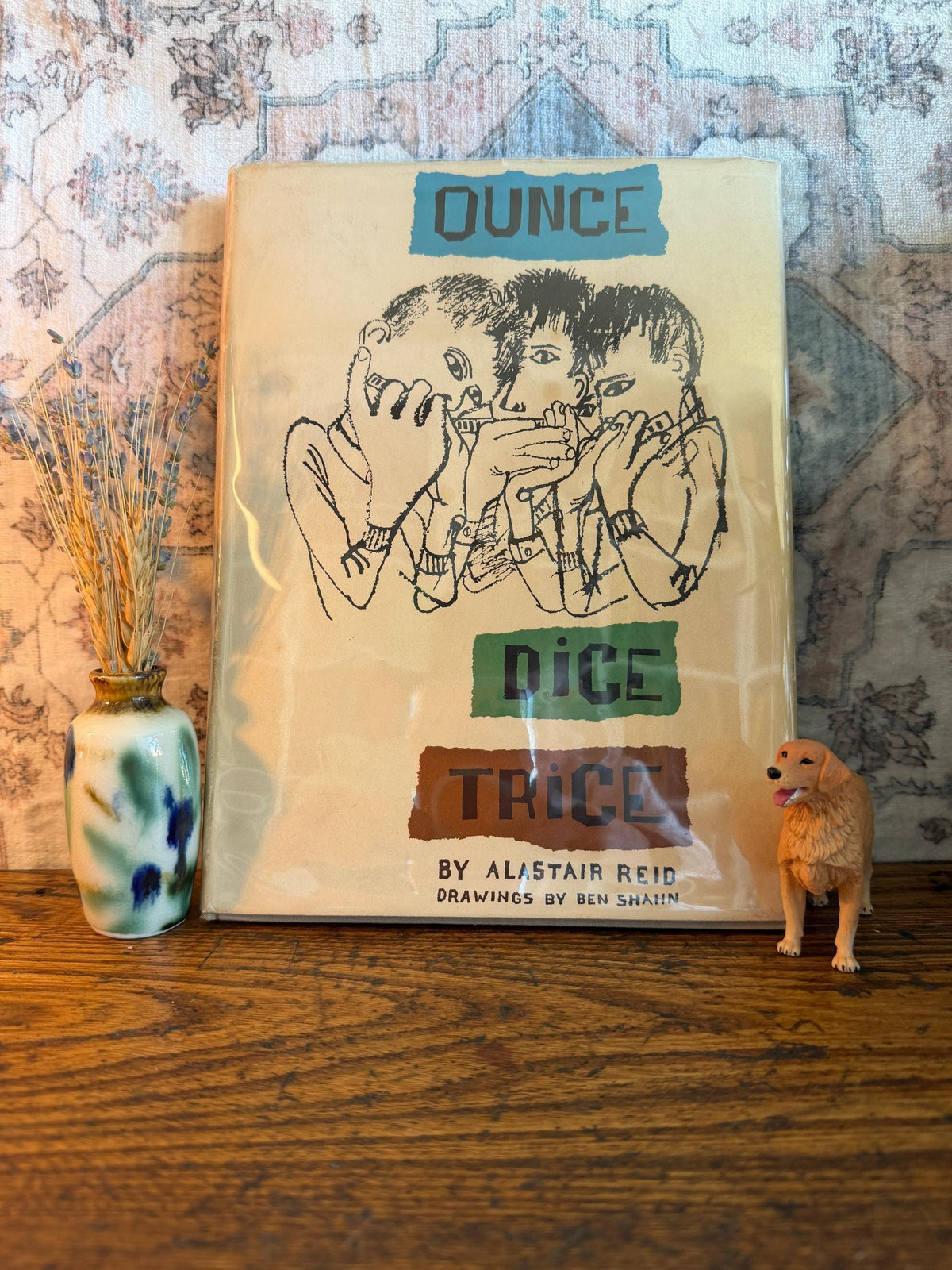 May include: A vintage book titled "Ounce Dice Trice" with illustrations of three figures. The cover is cream-colored with blue, green, and brown text. A small ceramic vase with wheat and a golden retriever figurine are next to the book.