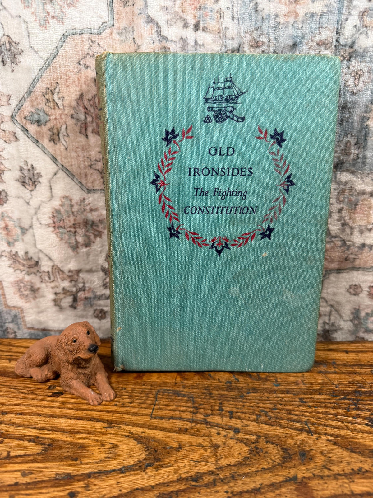 May include: A vintage book titled "Old Ironsides The Fighting Constitution" with a teal cover and decorative elements. A small brown dog figurine rests beside the book on a wooden surface.