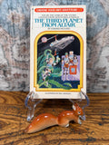 May include: A vintage paperback book titled "The Third Planet From Altair" with a colorful illustration of astronauts and a spaceship. A ceramic squirrel figurine rests in front of the book. The book is propped up on a clear plastic stand.