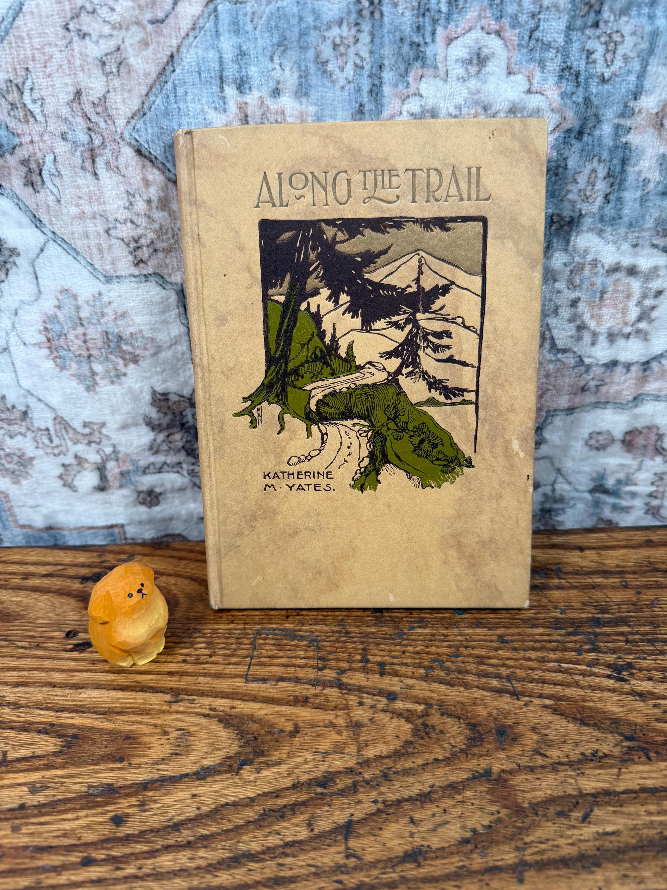 May include: A tan hardcover book titled Along the Trail by Katherine M. Yates, featuring a dark brown and green illustration of a mountain landscape, accompanied by a small, smooth, orange-yellow carved wooden figurine of a seated animal.
