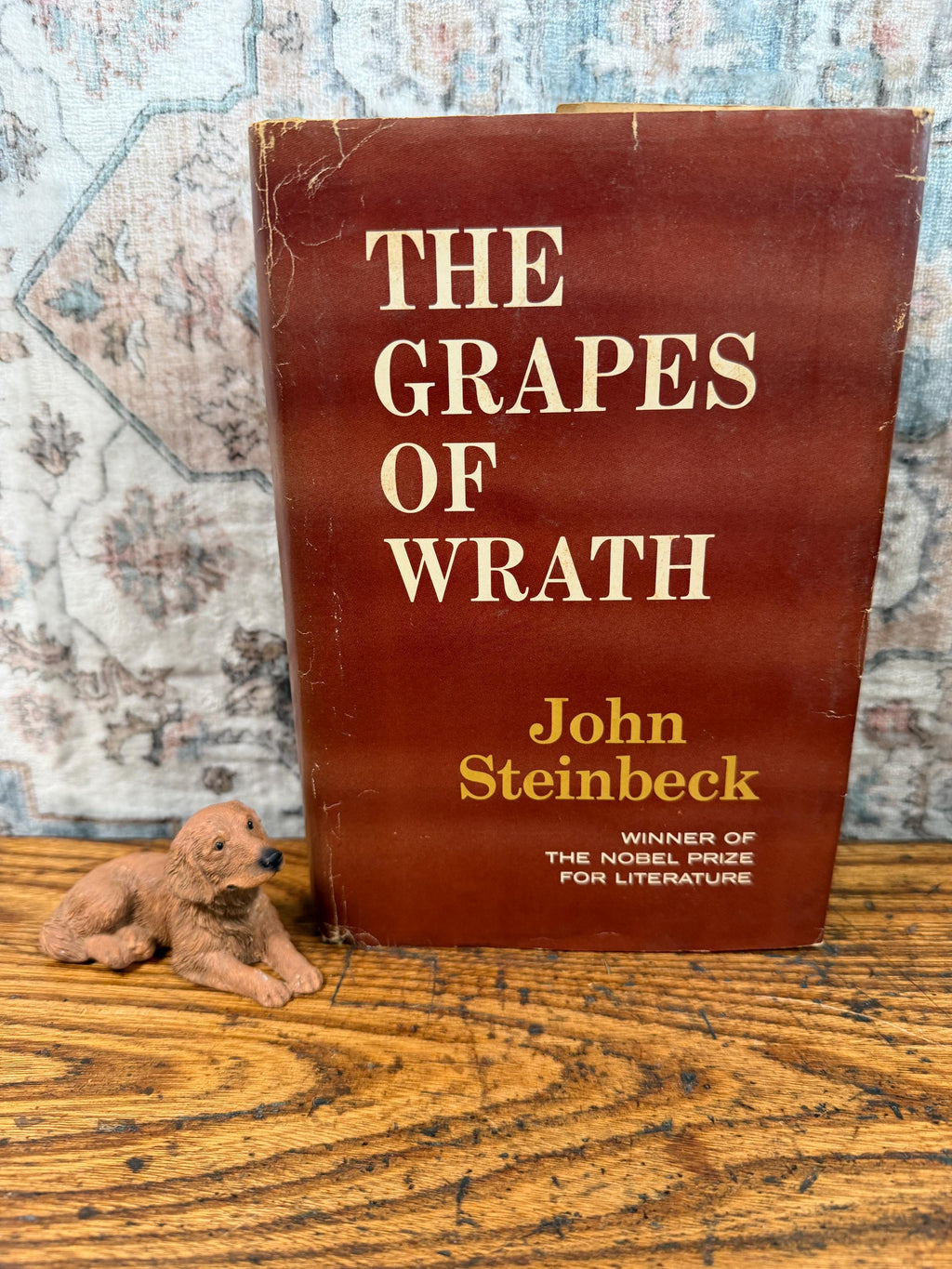 May include: A vintage copy of John Steinbeck's "The Grapes of Wrath" with a brown cover and white lettering. A small, brown dog figurine rests in front of the book on a wooden surface.