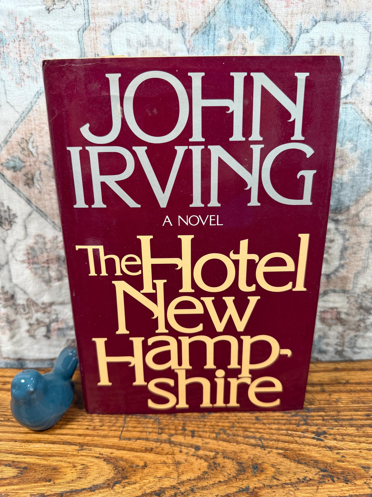 May include: A hardcover book titled "The Hotel New Hampshire" by John Irving. The book has a burgundy cover with white and gold lettering. A small blue ceramic bird figurine sits to the left of the book on a wooden surface.
