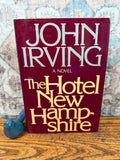 May include: A hardcover book titled "The Hotel New Hampshire" by John Irving. The book has a burgundy cover with white and gold lettering. A small blue ceramic bird figurine sits to the left of the book on a wooden surface.