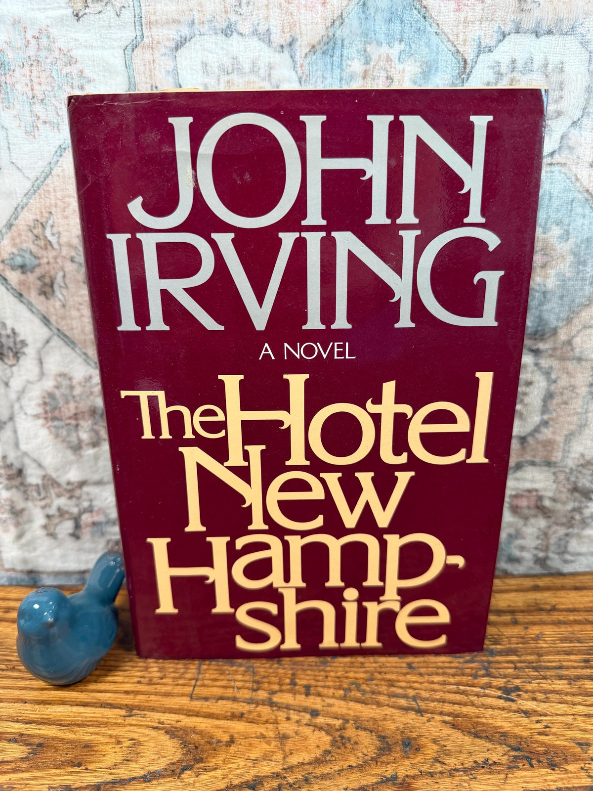 May include: A hardcover book titled "The Hotel New Hampshire" by John Irving. The book has a burgundy cover with white and gold lettering. A small blue ceramic bird figurine sits to the left of the book on a wooden surface.