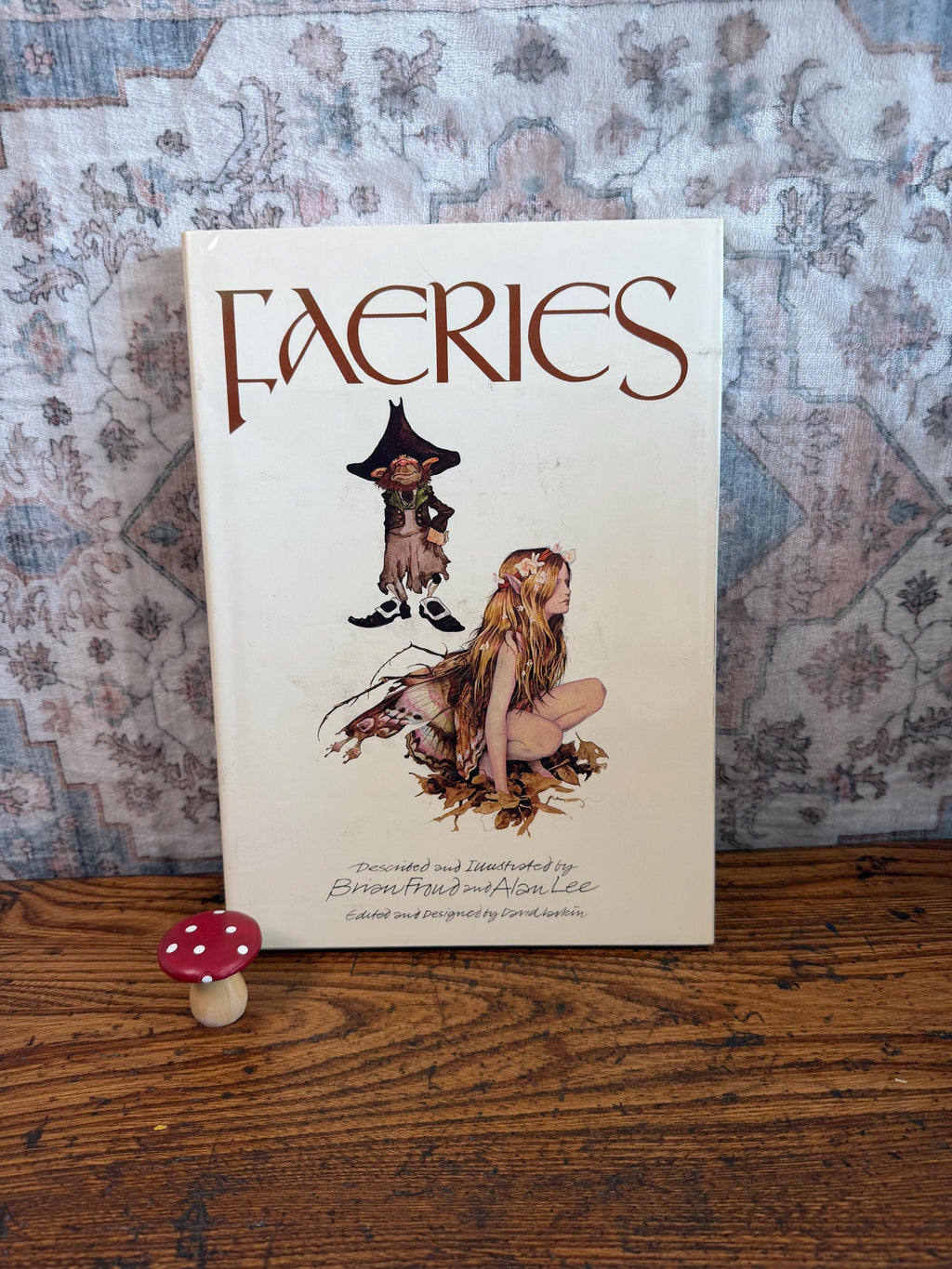 May include: A hardcover book titled Faeries, described and illustrated by Brian Froud and Alan Lee, featuring a central illustration of a small creature and a winged figure. A small red and white spotted mushroom figurine sits to the left.