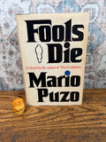 May include: A hardcover book titled Fools Die by Mario Puzo with a cream-colored cover, bold black lettering, and a small silhouette of a person in a coat. Beside the book sits a small, carved, light-brown wooden figurine of a seated dog.