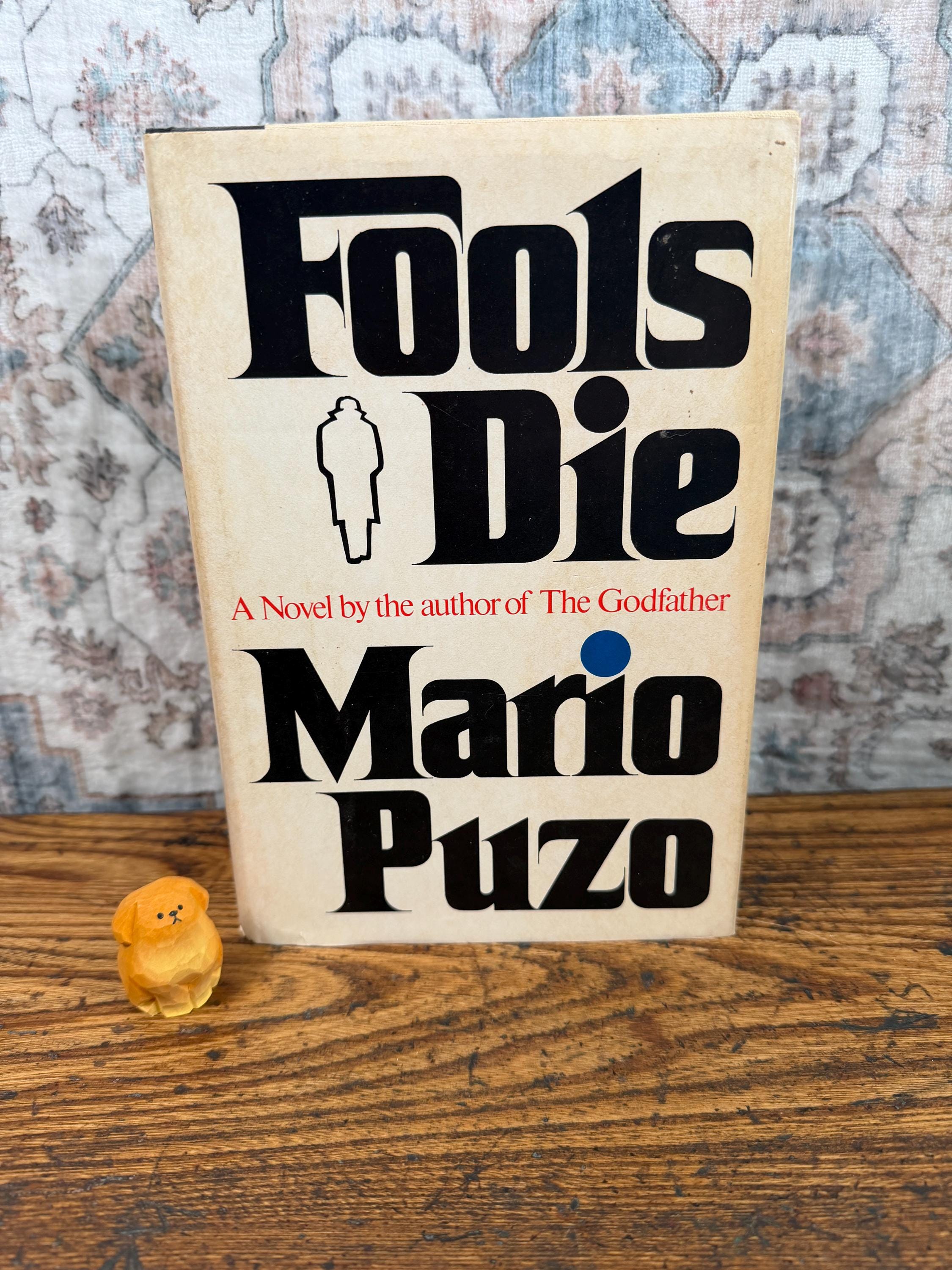 May include: A hardcover book titled Fools Die by Mario Puzo with a cream-colored cover, bold black lettering, and a small silhouette of a person in a coat. Beside the book sits a small, carved, light-brown wooden figurine of a seated dog.