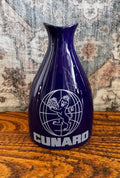 May include: A deep blue ceramic carafe with a narrow neck and a wide base. The front features a white graphic of a lion holding a globe inside a circular grid, with the word CUNARD printed in bold white capital letters underneath the emblem.