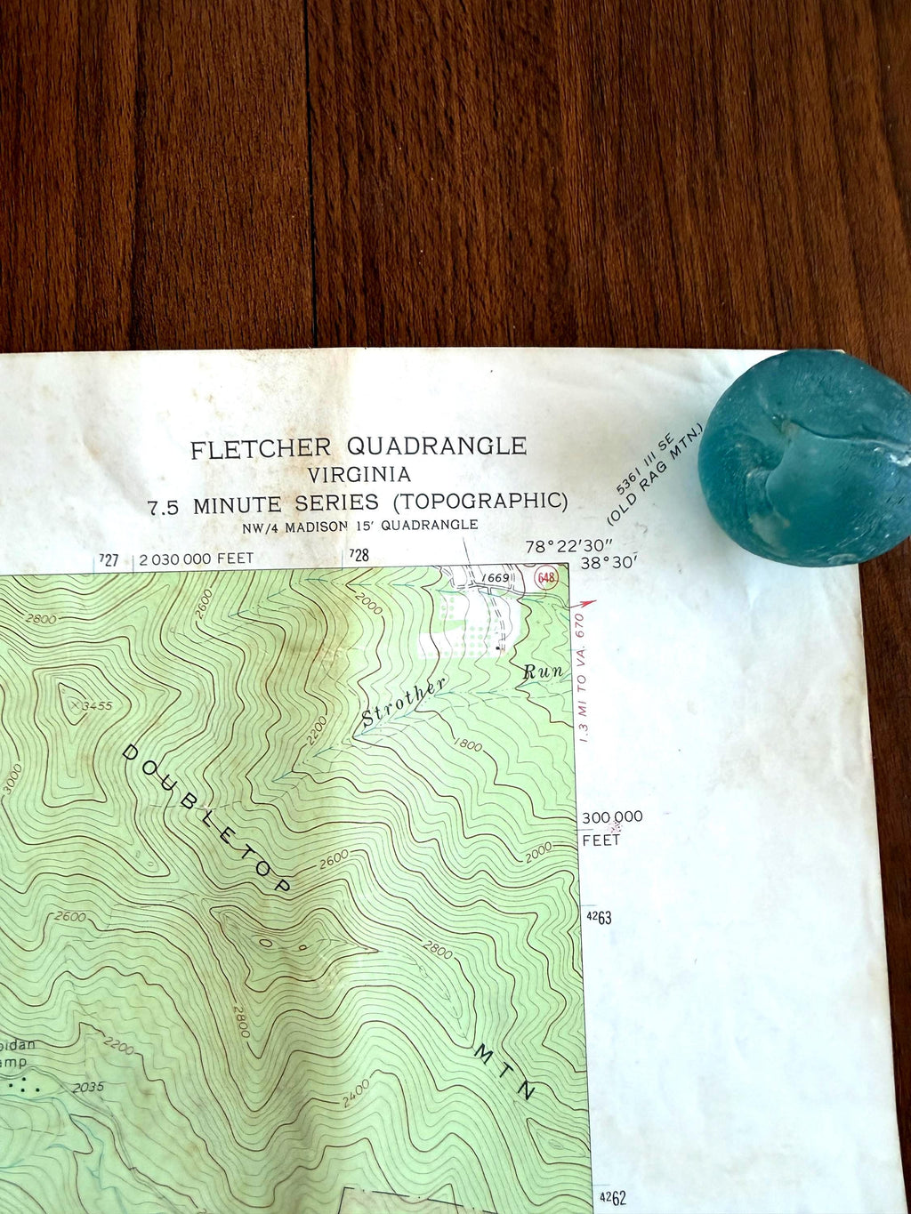 May include: A Fletcher Quadrangle Virginia 7.5 minute series topographic map featuring contour lines for Doubletop Mountain and Strother Run, with a round, translucent, light blue glass marble placed on the upper right corner of the paper.