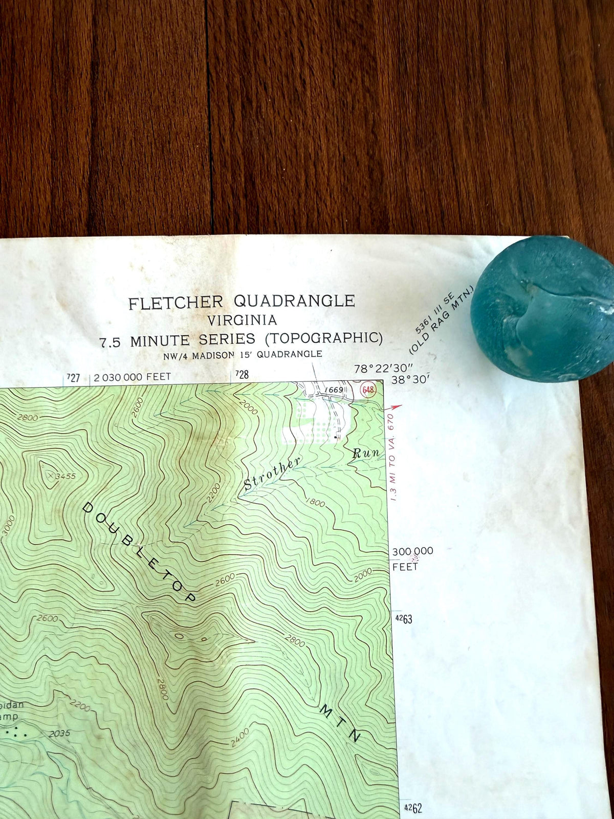 May include: A Fletcher Quadrangle Virginia 7.5 minute series topographic map featuring contour lines for Doubletop Mountain and Strother Run, with a round, translucent, light blue glass marble placed on the upper right corner of the paper.