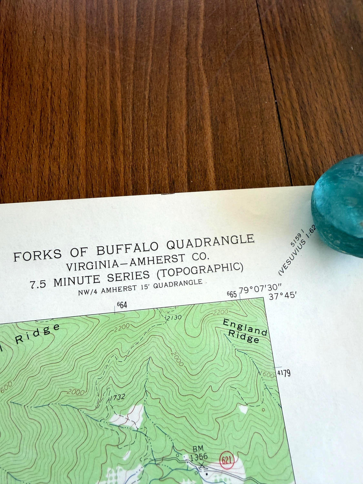 May include: A topographic map titled Forks of Buffalo Quadrangle Virginia-Amherst Co. 7.5 Minute Series, featuring green contour lines, elevation markers, and geographic coordinates, partially covered by a smooth, translucent teal glass stone.