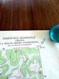 May include: A vintage Greenfield Quadrangle topographic map of Virginia, 7.5 minute series, featuring contour lines, road markers, and text indicating 2.9 miles to U.S. 250. A small, translucent, rounded teal glass object rests on the upper right corner.