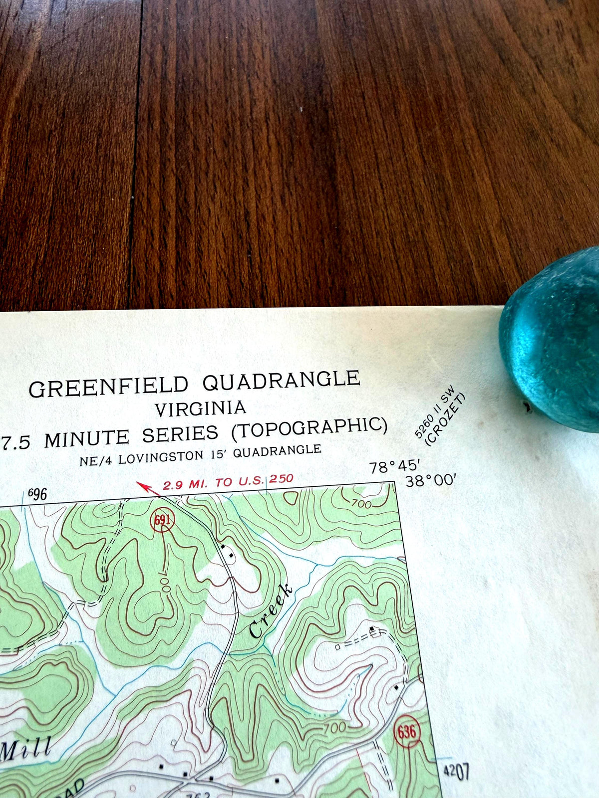 May include: A vintage Greenfield Quadrangle topographic map of Virginia, 7.5 minute series, featuring contour lines, road markers, and text indicating 2.9 miles to U.S. 250. A small, translucent, rounded teal glass object rests on the upper right corner.